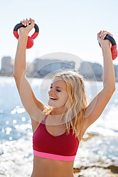 Happy young blonde woman pushing up kettlebells, outdoor workout