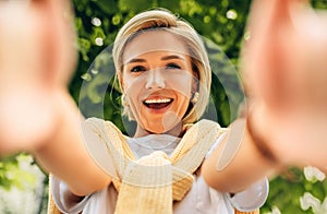 Happy young blonde woman smiling broadly making self portrait on her device outdoor. Happy pretty female taking selfie. People