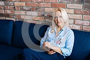 Happy young blonde woman using cell phone while sitting on soft blue sofa in living room