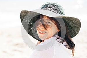 Happy young boho woman in hat relaxing and enjoying sunny warm day at ocean. Space for text. Stylish hipster girl sitting on beach