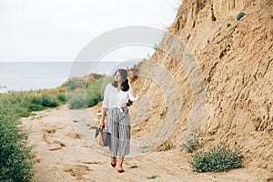 Happy young boho woman in white shirt walking on tropical island at sandy cliff and grass. Stylish hipster girl relaxing on beach