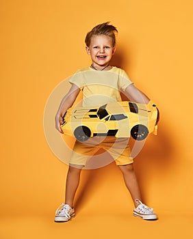 Happy young boy kid in yellow t-shirt and shorts holds a big huge yellow toy race car gift