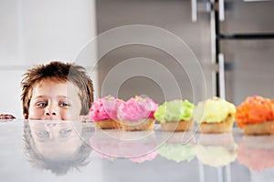 Happy young boy peaking over counter at row of cupcakes in kitchen