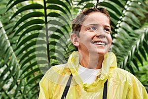 Happy young boy in raincoat standing in front of large fern during field trip