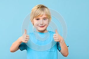 Happy young boy showing thumbs up and looking at camera isolated over pastel blue background. Cheerful kid.