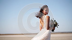 Happy young bride with bouquet of fresh flowers going at desert landscape. Elegance lady in gorgeous wedding dress walks