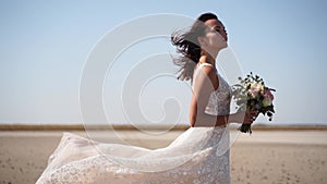 Happy young bride with bouquet of fresh flowers going at the dry lake. Elegance lady in gorgeous wedding dress walks in