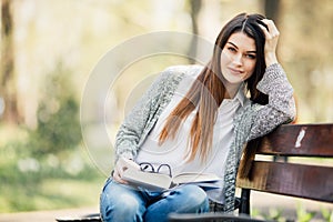 Happy young brunette with a notebook in hands sitting on a park bench