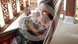 Happy young brunette woman relaxing in a hammock with Maine Coon cat at garden in sunny summer day, slow motion