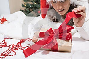 Happy young brunette woman in santa claus hat opens a gift box for Christmas lying on a white bed. Christmas gifts.