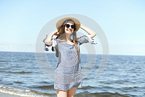 Happy young brunette woman standing on the ocean beach and looking at camera while smiling, wearing fashion hat and