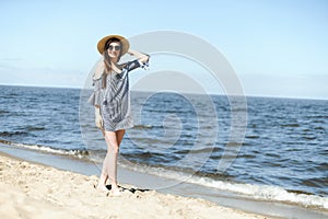 Happy young brunette woman standing on the ocean beach and looking at camera while smiling, wearing fashion hat and