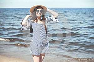 Happy young brunette woman standing on the ocean beach and looking at camera while smiling, wearing fashion hat and