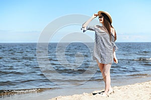 Happy young brunette woman standing on the ocean beach while smiling, and wearing fashion hat, sunglasses. The enjoying