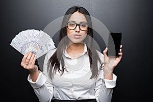 Happy young brunette woman in white shirt showing smartphone with blank screen and cash money in hands on black backgroun