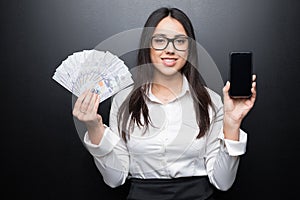 Happy young brunette woman in white shirt showing smartphone with blank screen and cash money in hands on black backgroun