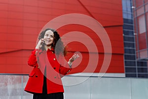Happy young brunette young woman, wear in red, talking on mobile phone and gesture with hand, over building background.