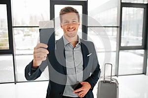 Happy young buisnessman stand in airport hall and show passport with ticket. He looks on camera and smile. Guy has phone