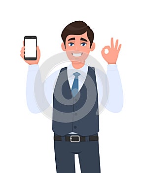 Happy young business man in vest suit showing a blank screen mobile phone and gesturing okay, OK sign. Person holding smartphone.