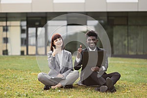 Happy business people, sitting on green grass in front of office, showing success with thumb up