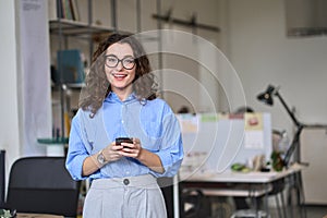 Happy young business woman holding phone standing in office using mobile.