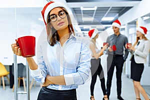 Happy young business woman in Santa Claus hat, holding a red mug, with a group of colleagues in the background, during a coffee