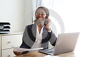 Happy young business woman sitting at office desk talking on mobile phone with a document in hand