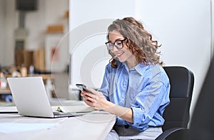 Happy young business woman using cellphone working in office sitting at desk.