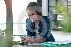 Happy young business woman using her mobile phone while working with computer in the kitchen at home