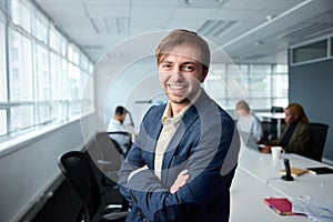 Happy young businessman in businesswear with arms crossed next to desk in office