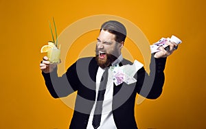 Happy young businessman with glass of cocktail in formal clothes holding bunch of money banknotes