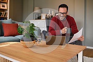 Happy young businessman holding coffee cup and document discussing over video call on laptop at home