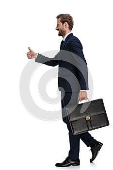 Happy young businessman holding suitcase and making thumbs up sign