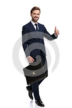 Happy young businessman holding suitcase and making thumbs up sign