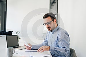 Happy young businessman looking at papers graphs and charts using laptop at his office desk