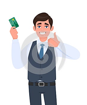 Happy young businessman showing credit, debit card. Man making or gesturing thumbs up sign. Successful person in waistcoat holding