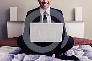 Happy young businessman sitting in bed with laptop