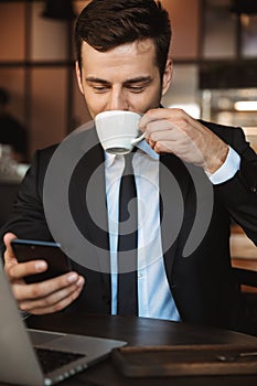 Happy young businessman sitting in cafe using laptop computer drinking coffee using mobile phone