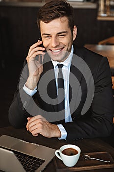 Happy young businessman sitting in cafe using laptop computer talking by mobile phone
