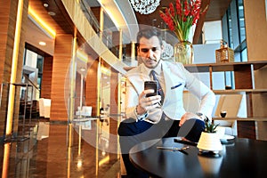 Happy young businessman sitting relaxed on sofa at hotel lobby using smartphone