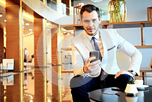 Happy young businessman sitting relaxed on sofa at hotel lobby using smartphone