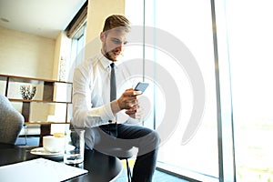 Happy young businessman sitting relaxed on sofa at hotel lobby using smartphone, waiting for someone