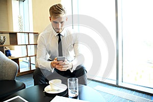 Happy young businessman sitting relaxed on sofa at hotel lobby using smartphone, waiting for someone