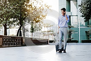 Happy Young Businessman Using Mobile Phone while Walking with Suitcase in the City. Lifestyle of Modern People