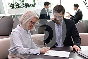 Happy young businessman watching smiling mature businesswoman signing agreement.