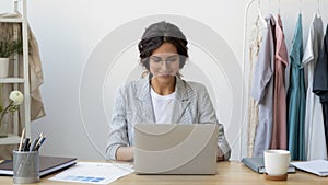 Happy young businesswoman self-employed tailor sitting at desk using laptop