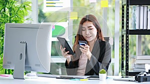 Happy young businesswoman sitting in her office holding a credit card, looking at digital tablet, and provide her data to make an