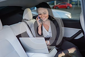 Happy young businesswoman typing at laptop and talking on smartphone while sitting on the back seat of car. Working