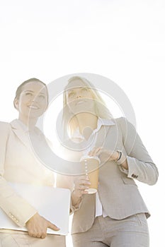 Happy young businesswomen with disposable cup and laptop against sky