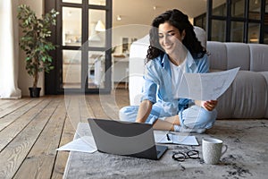 Happy young Cauasian lady working or studying on laptop computer, holding documents, looking at pc screen indoors photo